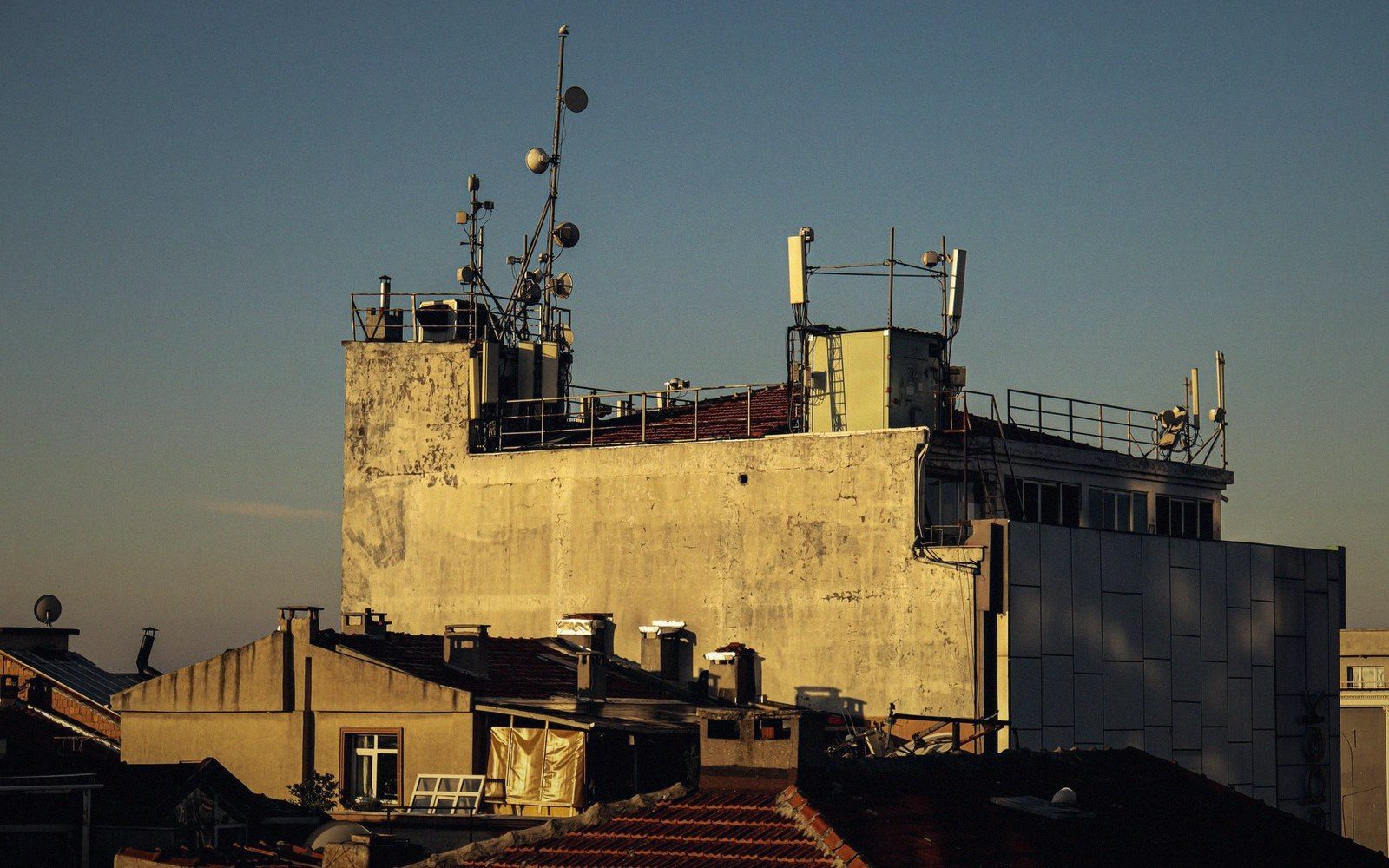 Antennas and satellite dishes cluster atop a weathered building above tiled rooftops
