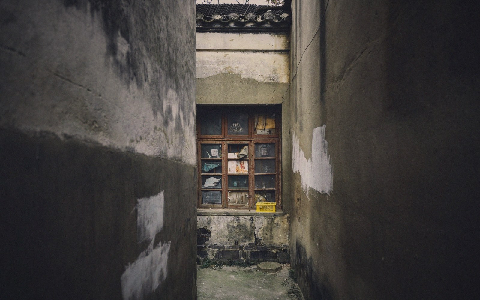 A narrow alley between weathered walls leads to an old wooden window with a yellow crate below