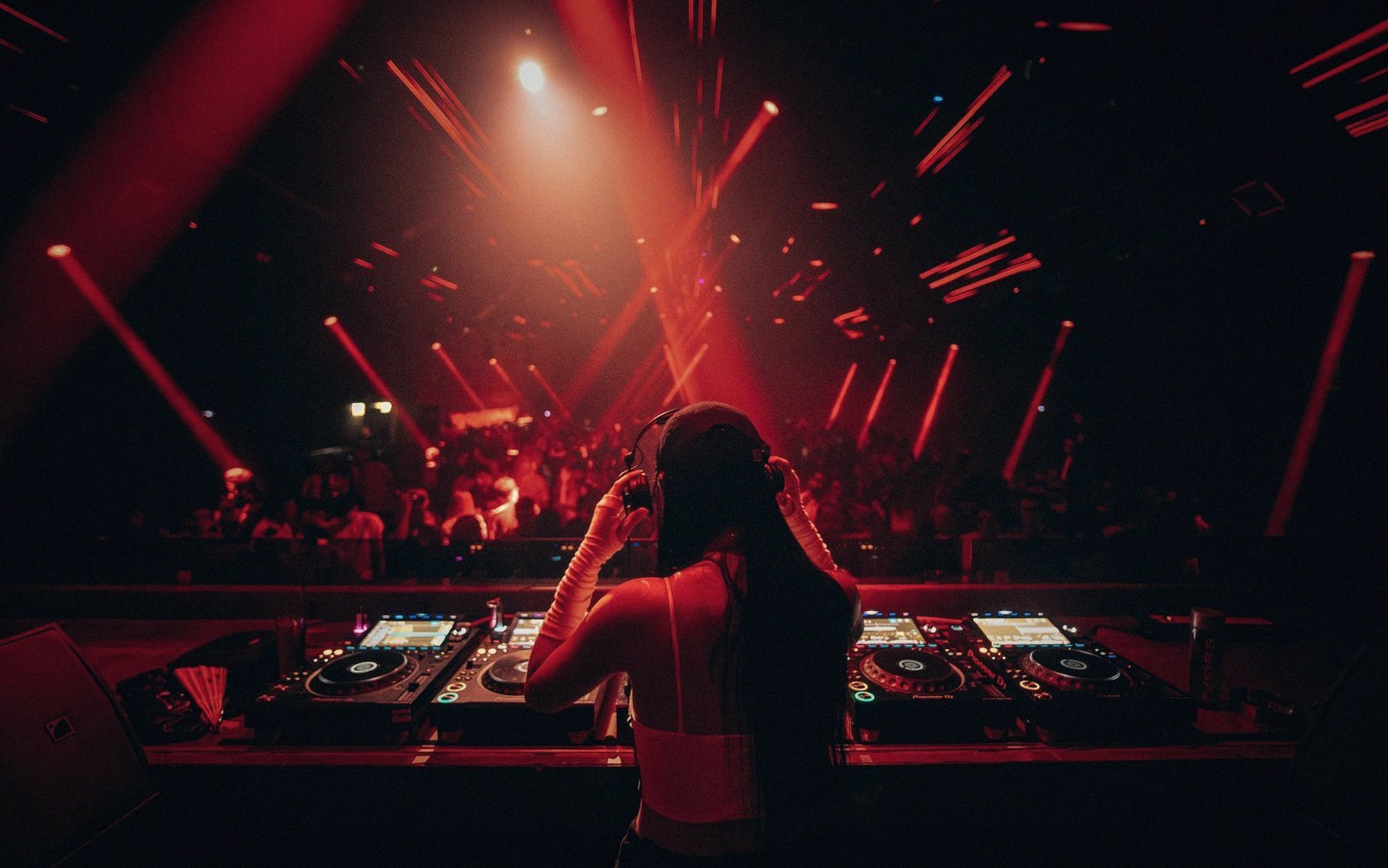 A DJ wearing headphones performs at a mixing deck under red laser lights over a crowd